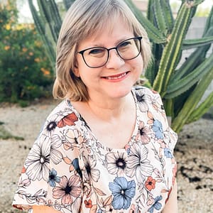 Smiling Sue Hegg with short gray hair and black glasses in a floral blouse, outdoors with tall cacti in the background.
