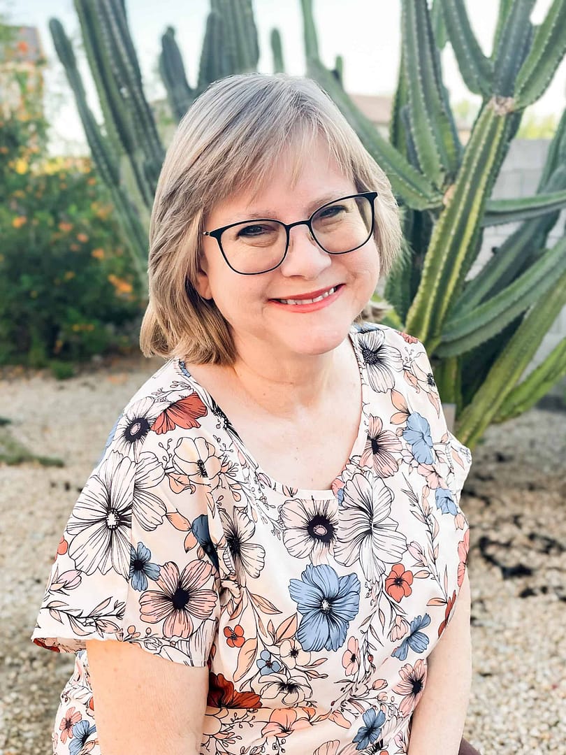 Smiling Sue Hegg with short gray hair and black glasses in a floral blouse, outdoors with tall cacti in the background.